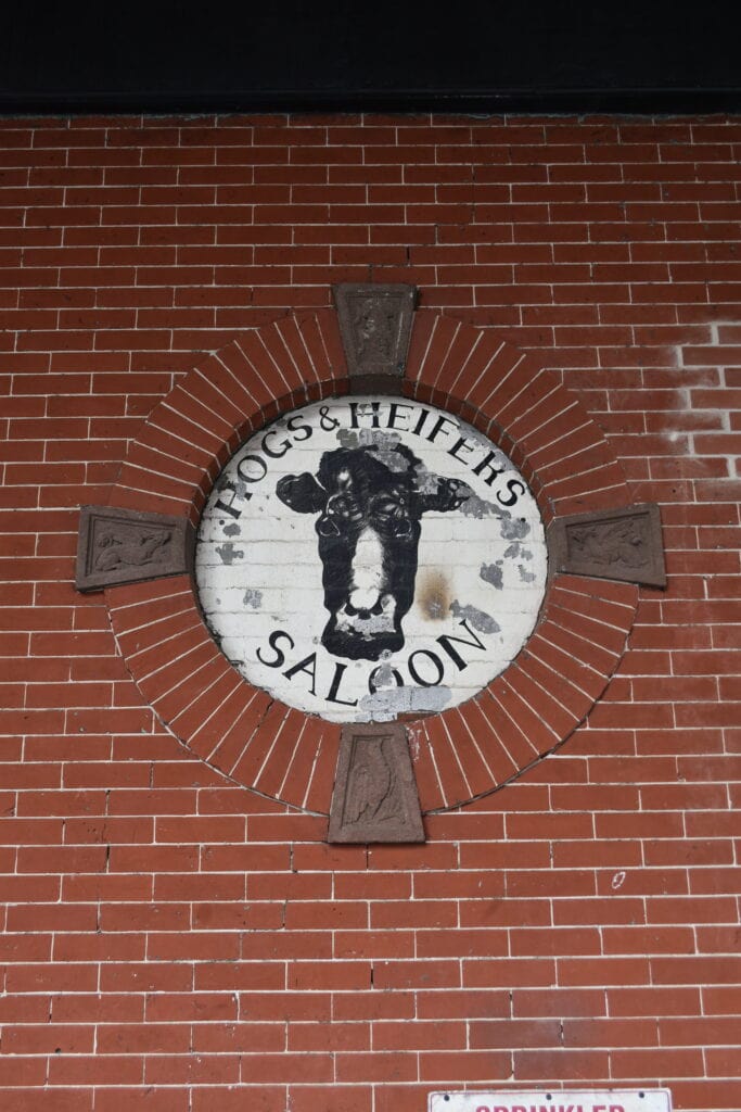 Circular sign on brick wall reads "Hogs & Heifers Saloon" with a cow head illustration in the centre, marking a classic spot in one of the neighbourhood’s in New York.