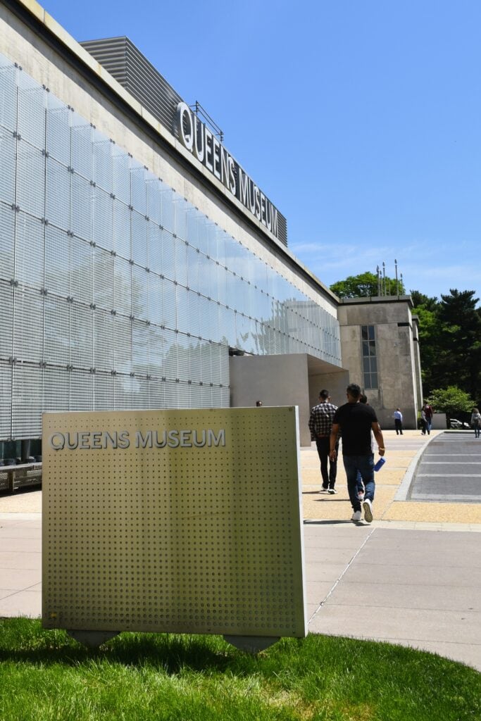 People stroll by the glass exterior of the Queens Museum on a sunny day, passing a sign in front, enjoying one of the vibrant parks in New York.