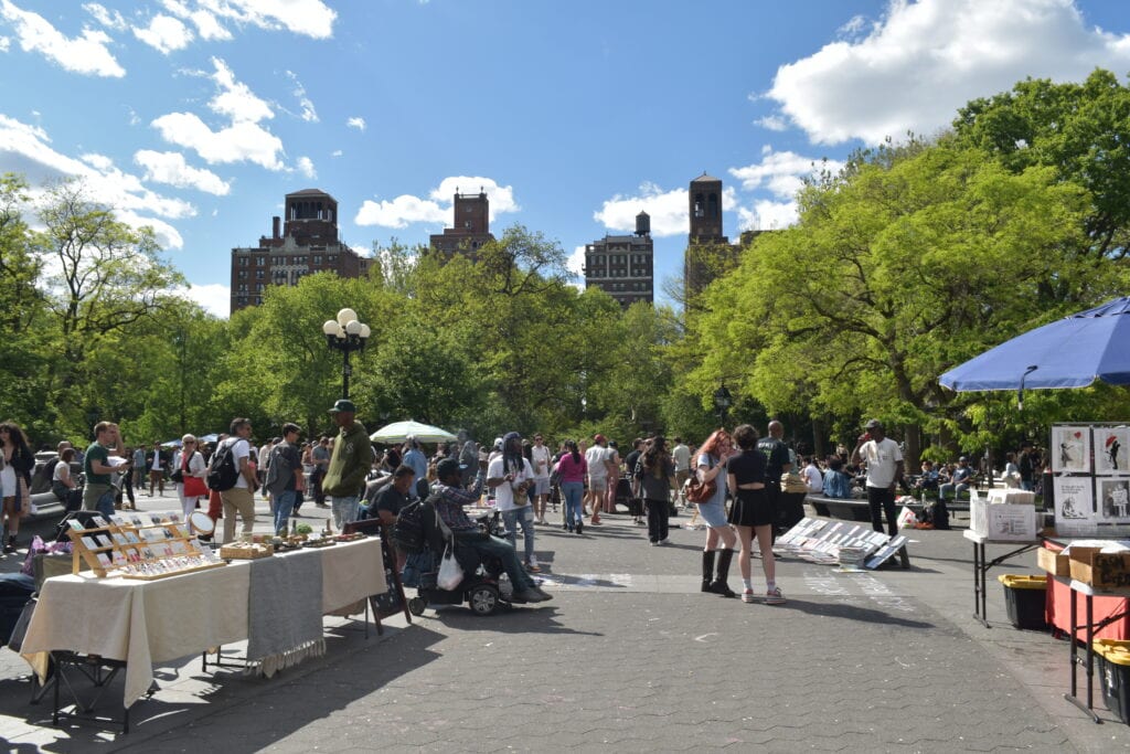 People browse art and craft stalls at a busy outdoor market in a sunny park, surrounded by tall buildings in one of the neighbourhood’s in New York.