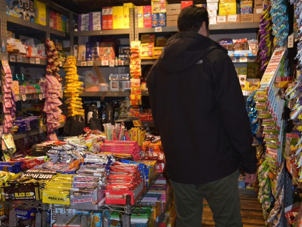 Person in a black jacket browsing a well-stocked candy and snack aisle in a small New York store, seeking insider tips on the best local treats.