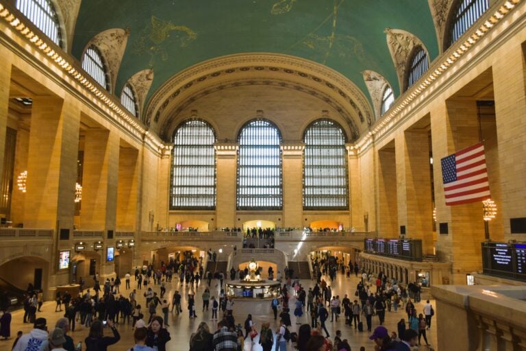 Crowds gather under arched windows and a teal ceiling in Grand Central station’s main concourse.