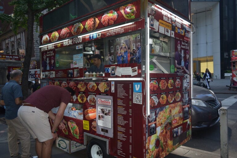 A man orders food at a street stall covered in colourful images of various halal dishes on a busy New York city pavement, capturing the vibrant spirit of street food in New York.