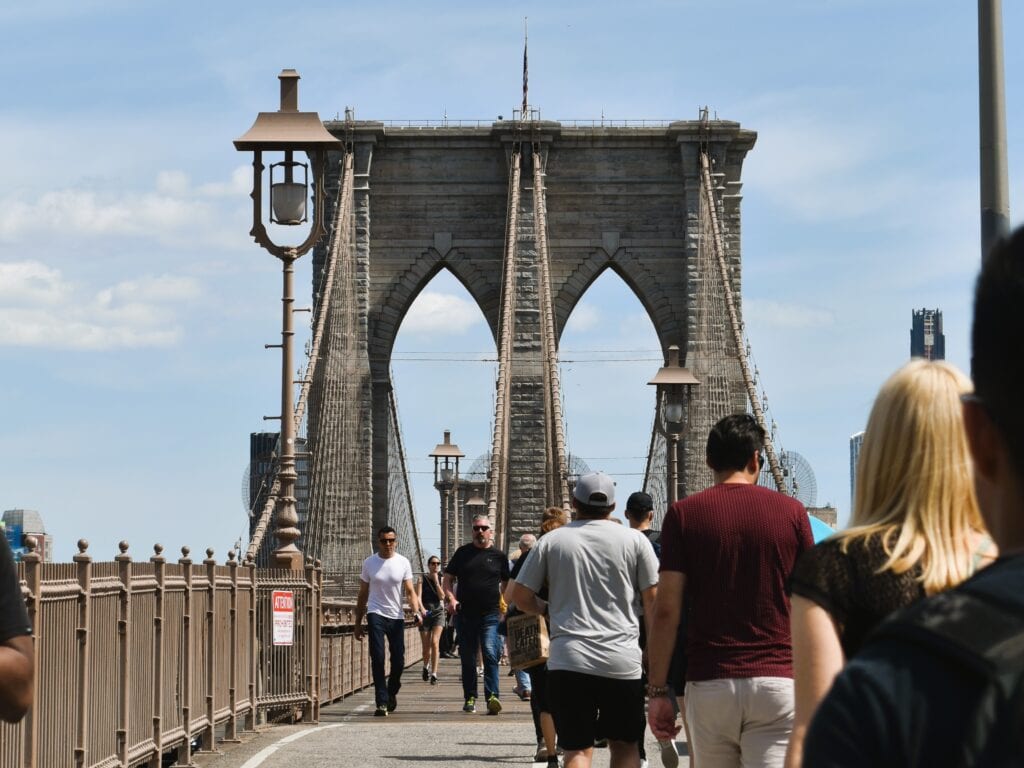People walking on the Brooklyn Bridge pedestrian path under a clear blue sky, with the iconic Brooklyn Bridge towers rising ahead.