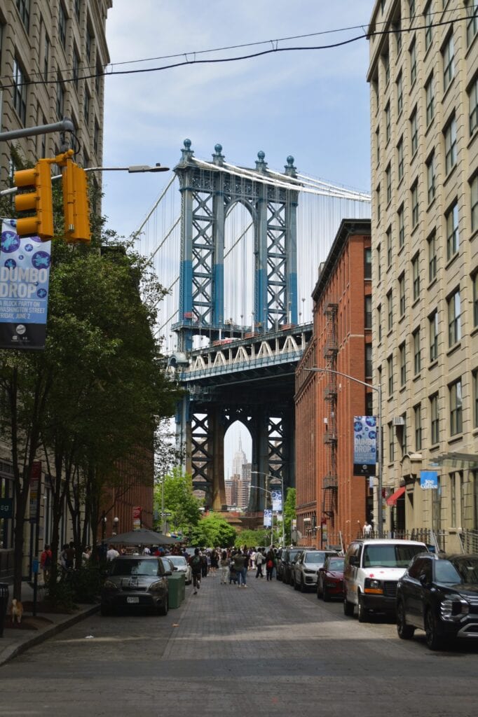 View of Manhattan Bridge framed by buildings in DUMBO, Brooklyn, with people and cars on the street, just a short walk from the iconic Brooklyn Bridge.