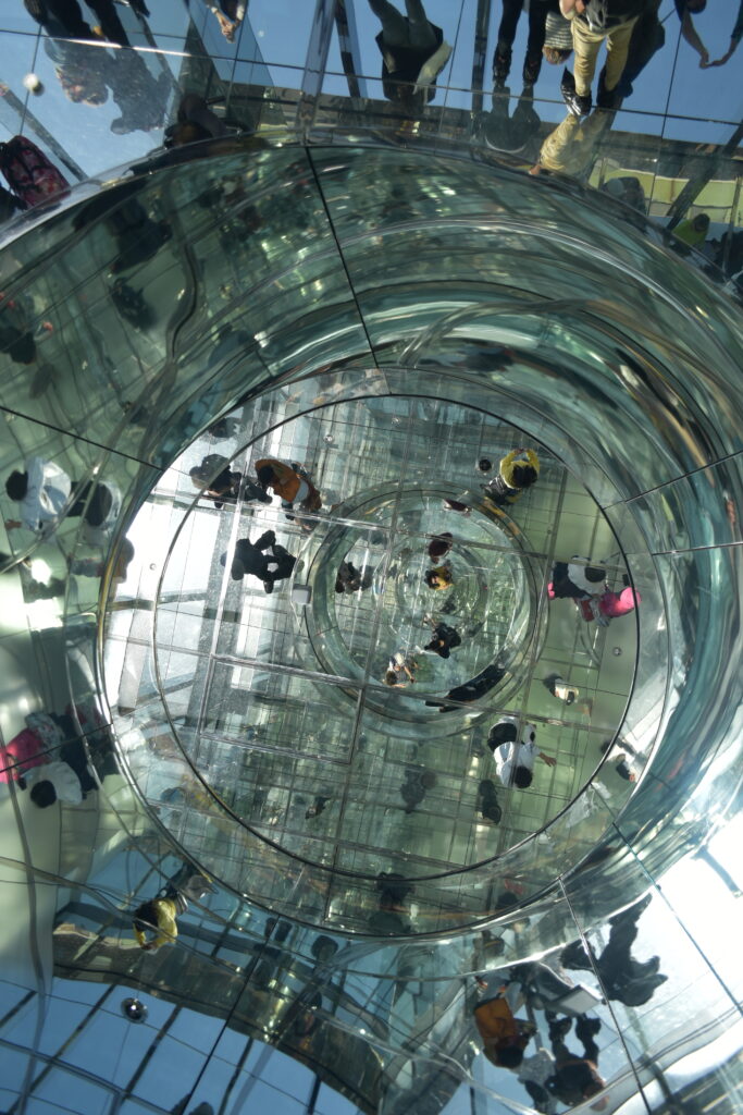 People reflected in a large, mirrored, spiral structure, creating a dizzying and abstract visual effect—one of the best skyviews in New York for capturing unique city reflections.