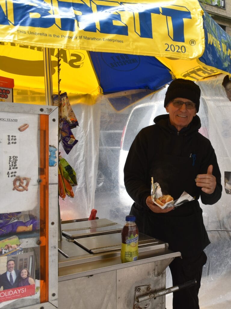 A smiling street vendor in a black beanie serves food you need to try in New York at a hot dog stall under a yellow umbrella.
