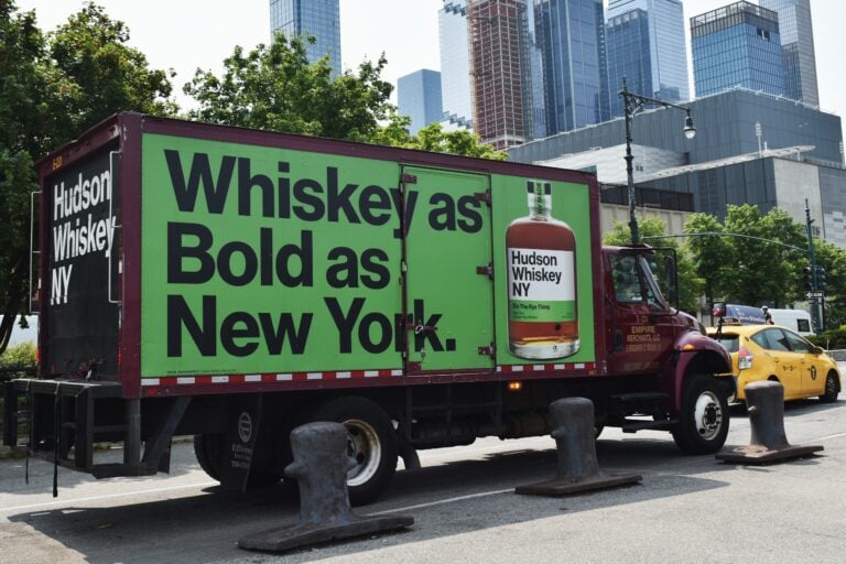 A truck with an ad for Hudson Whiskey NY, reading "Whiskey as Bold as New York," parked on a city street, offers New York insider tips to those who appreciate the city's bold spirit.