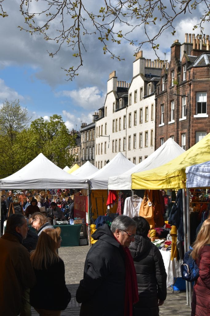 People browse stalls with white canopies at an outdoor market, beside historic buildings on a sunny day, experiencing one of the best attractions Edinburgh has to offer.