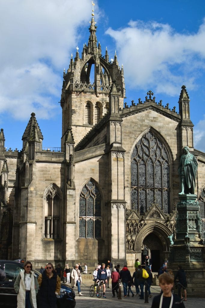 People walk in front of a large historic stone church with tall stained glass windows and a pointed tower, one of the best attractions Edinburgh has to offer.