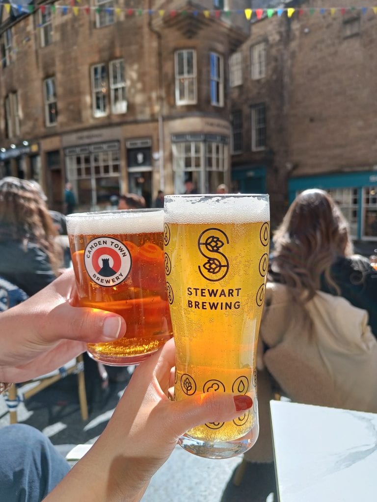 Two people toast with Stewart Brewing and Camden Town Brewery beers at an outdoor café on a sunny day, soaking in the lively atmosphere after exploring the best attractions Edinburgh has to offer.
