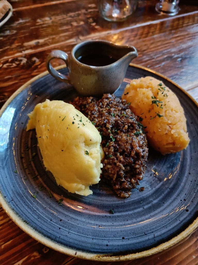 A plate with mashed potatoes, haggis, neeps, and a small jug of brown gravy on a wooden table—a classic taste of Scotland often enjoyed after exploring the best attractions Edinburgh has to offer.