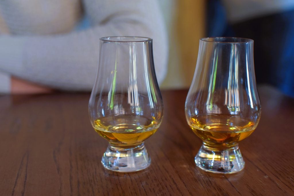 Two glasses of amber-colored whiskey on a wooden table, with a person blurred in the background—perfect after exploring the best attractions Edinburgh has to offer.