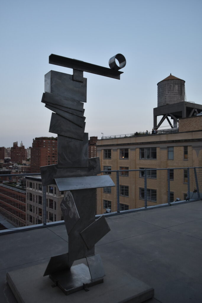 A metallic abstract sculpture graces a rooftop, overlooking museums in New York and a cityscape bathed in the soft glow of dusk, with a water tower standing sentinel.