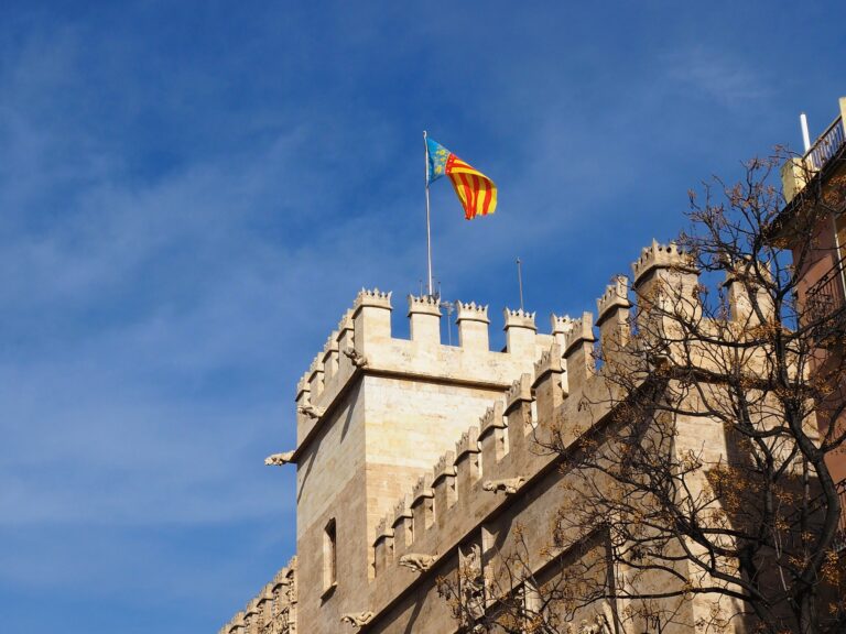 Stone castle with battlements and a red and yellow striped flag atop, under a clear blue sky in Valencia, perfect for celebrating festive occasions.