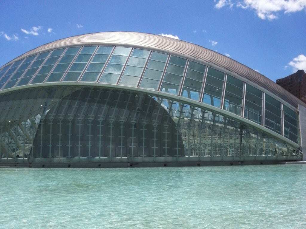 A futuristic glass and steel building, reminiscent of the Ciutat de les Arts i les Ciències, is reflected in clear blue water under a sunny sky.