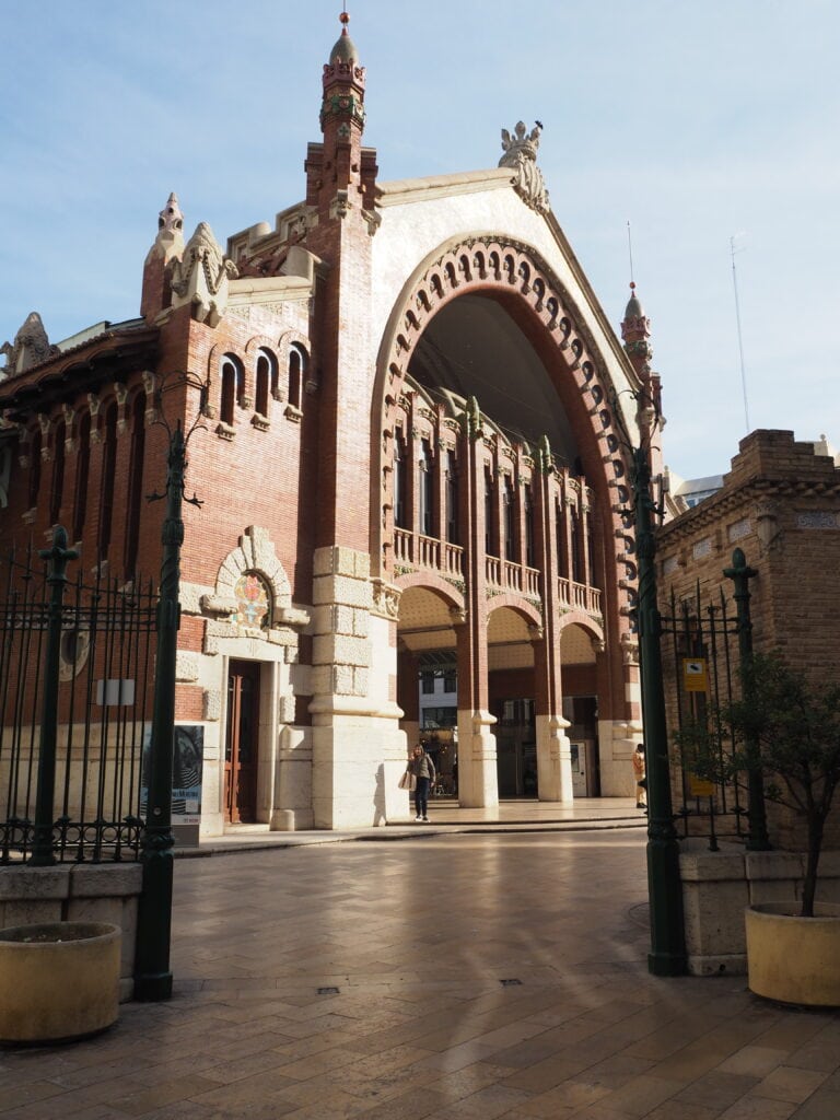 Historic brick market building with a large arched entrance and ornate architectural details, featured among the 7x Best things to Do in Valencia, set in a sunlit square.