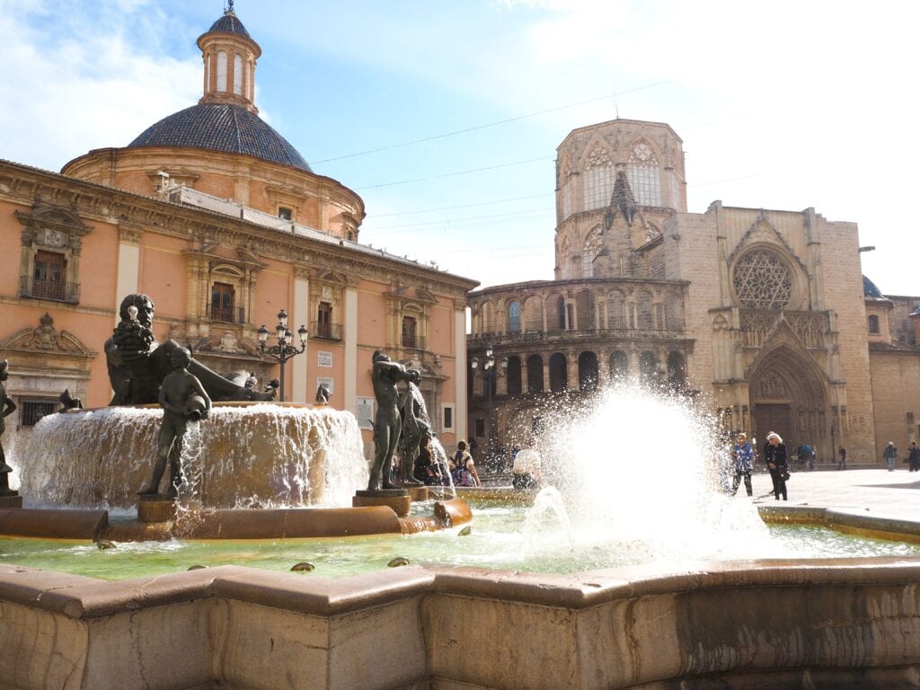 A historic square with a fountain and statues, surrounded by old buildings and a cathedral under a bright sky—one of the 7x Best things to Do in Valencia.