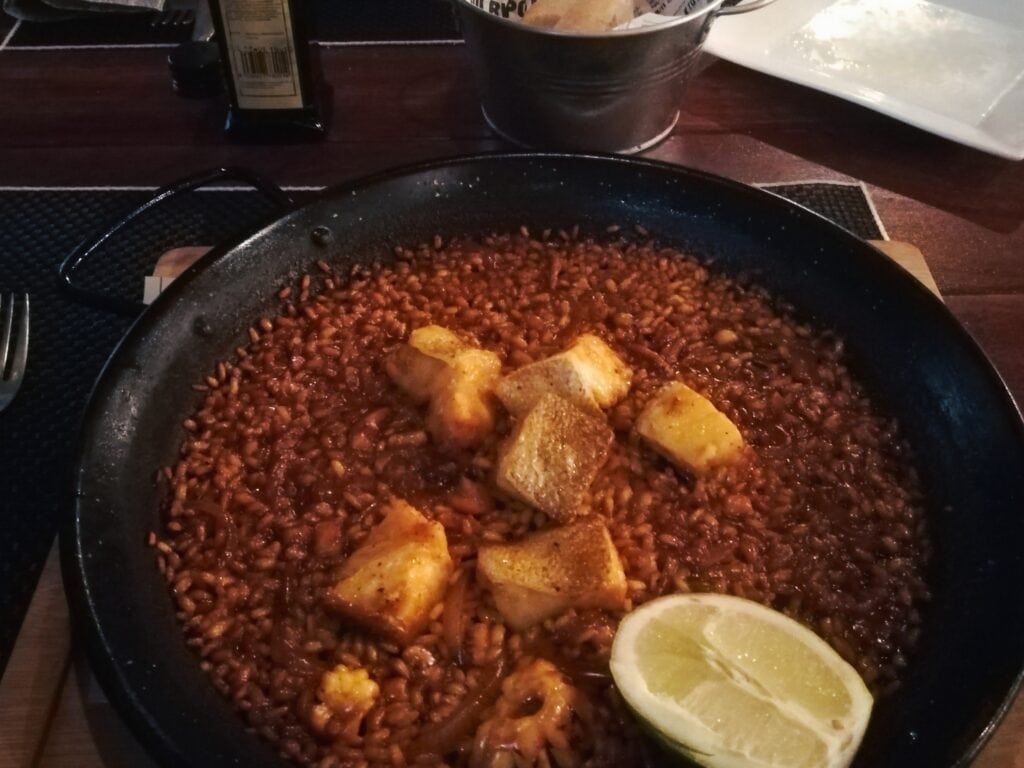 A pan of cooked rice with fish pieces and a lime wedge, served on a dark table—an inviting example of Spanish Food and Drink you need to try.