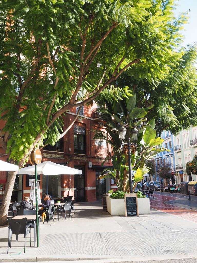 Sunny outdoor café with tables and umbrellas under leafy trees on a city street, nestled among colorful buildings in one of the Popular Neighborhoods in Valencia.