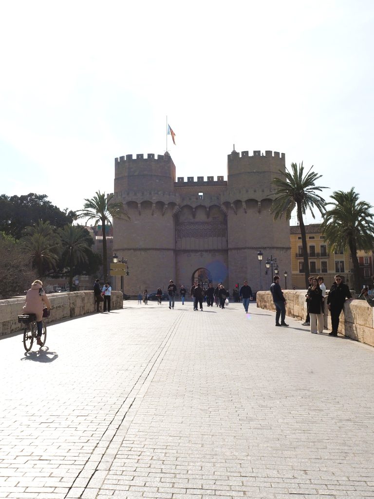 People walk and cycle toward a historic stone gate with towers, palm trees, and a flag—an iconic sight in one of the Popular Neighborhoods in Valencia.