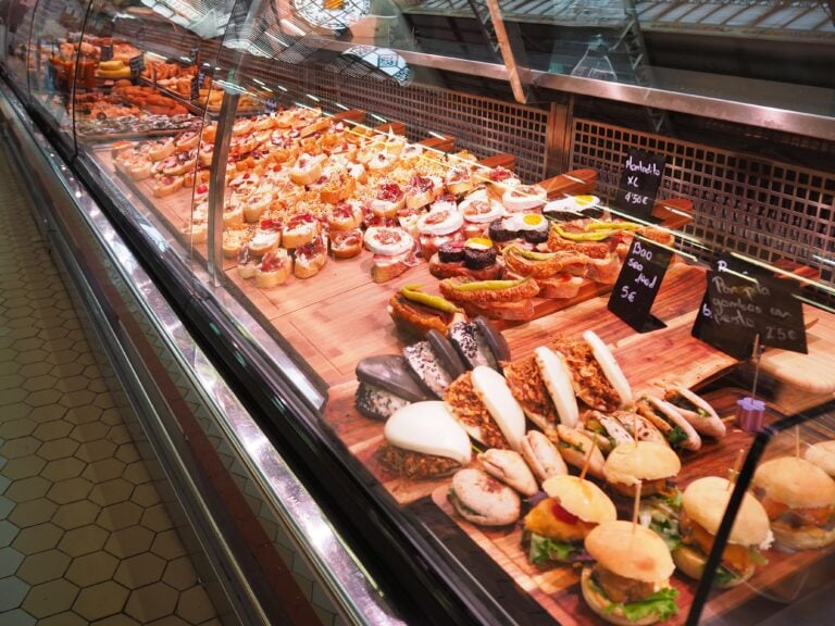 A deli counter displays assorted sandwiches, pastries, and snacks with price tags on a wooden board, featuring tempting Spanish food and drinks you need to try.
