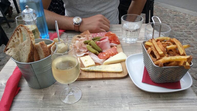 A table with wine, bread, cheese, cold cuts, pickles, and a basket of chips evokes the charm of the best wine tastings near Avignon, with a person seated behind.