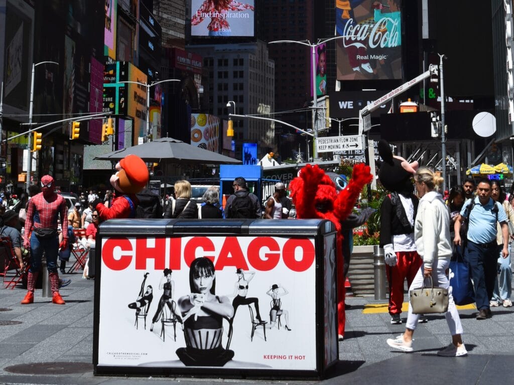Times Square scene with a "CHICAGO" billboard, West End Musicals vibes, costumed characters, and people walking on a sunny day.