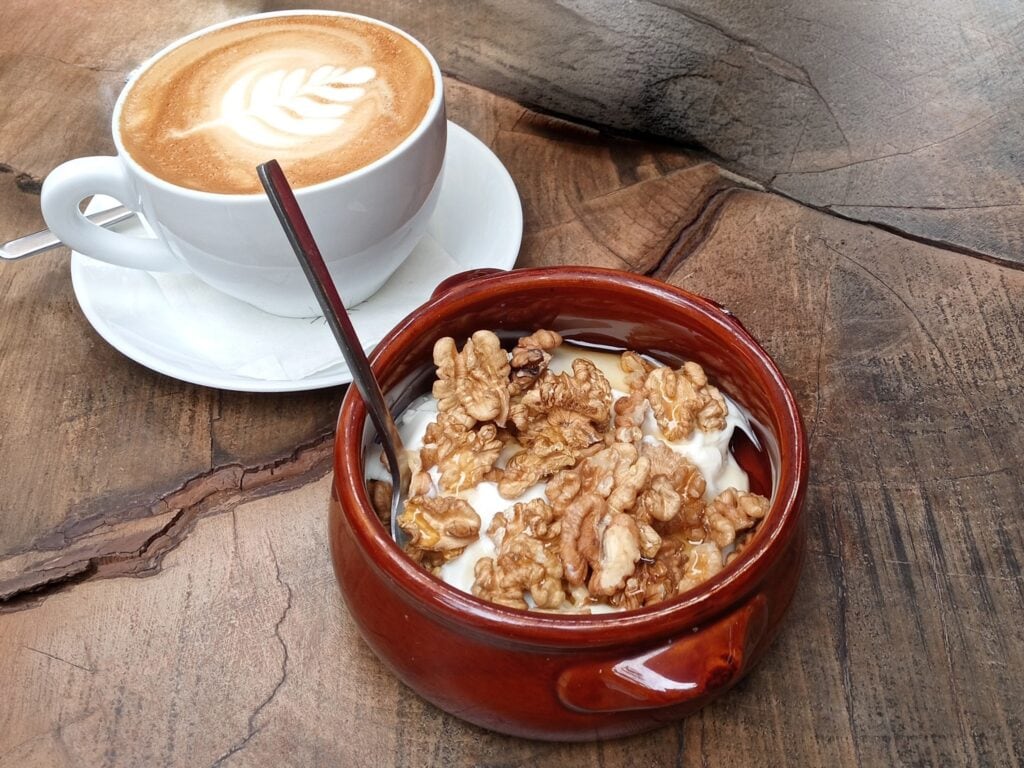 A cup of latte with latte art and a bowl of yoghurt topped with walnuts on a rustic wooden table—an example of the best comfort food Athens has to offer.