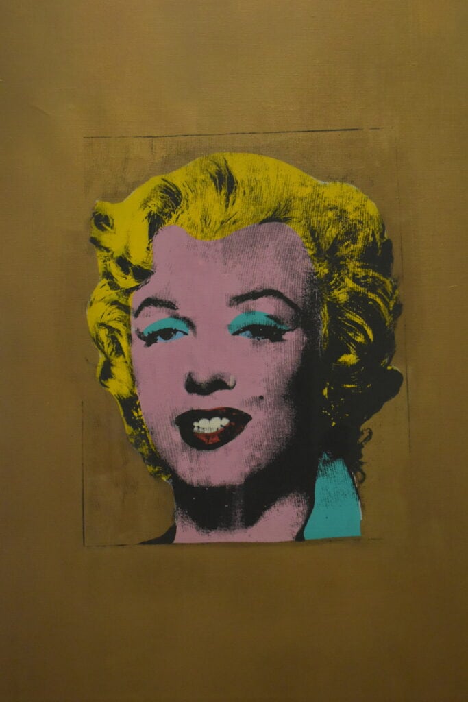 A pop art portrait of a woman with yellow hair, pink skin, blue eyeshadow, and red lips on a gold background—just the kind of iconic artwork you might see when you visit MoMA in New York (Museum of Modern Art).