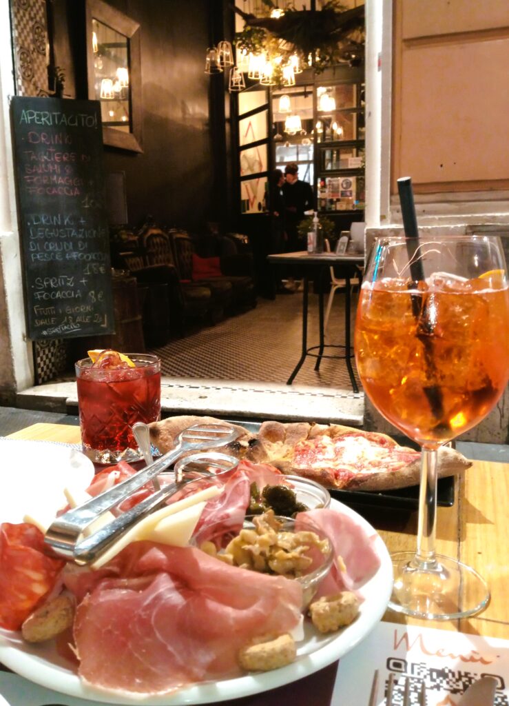 A cozy restaurant perfect for aperitivo time in Rome, featuring a charcuterie plate, cocktails, and pizza on the table, with a chalkboard menu in the background.