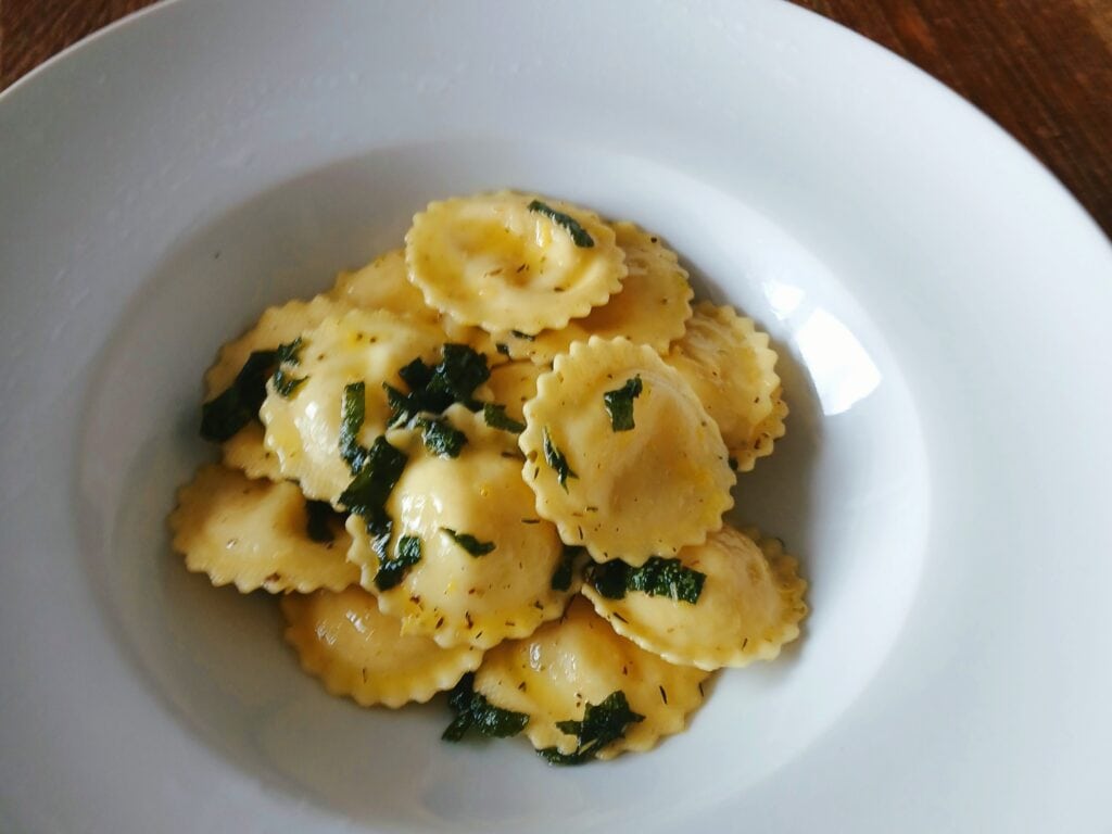 A white bowl filled with ravioli pasta, one of the 6x best Italian dishes, topped with chopped herbs and a light sauce.