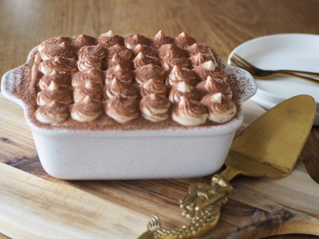 A dish of tiramisu dusted with cocoa powder on a wooden board, with plates and cutlery nearby—one of the 6x best Italian dishes to savour.