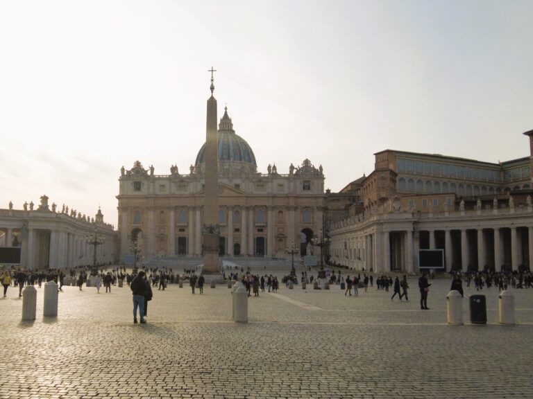 St. Peter's Square with tourists walking and St. Peter's Basilica in the background under a clear sky in Vatican City, Rome.