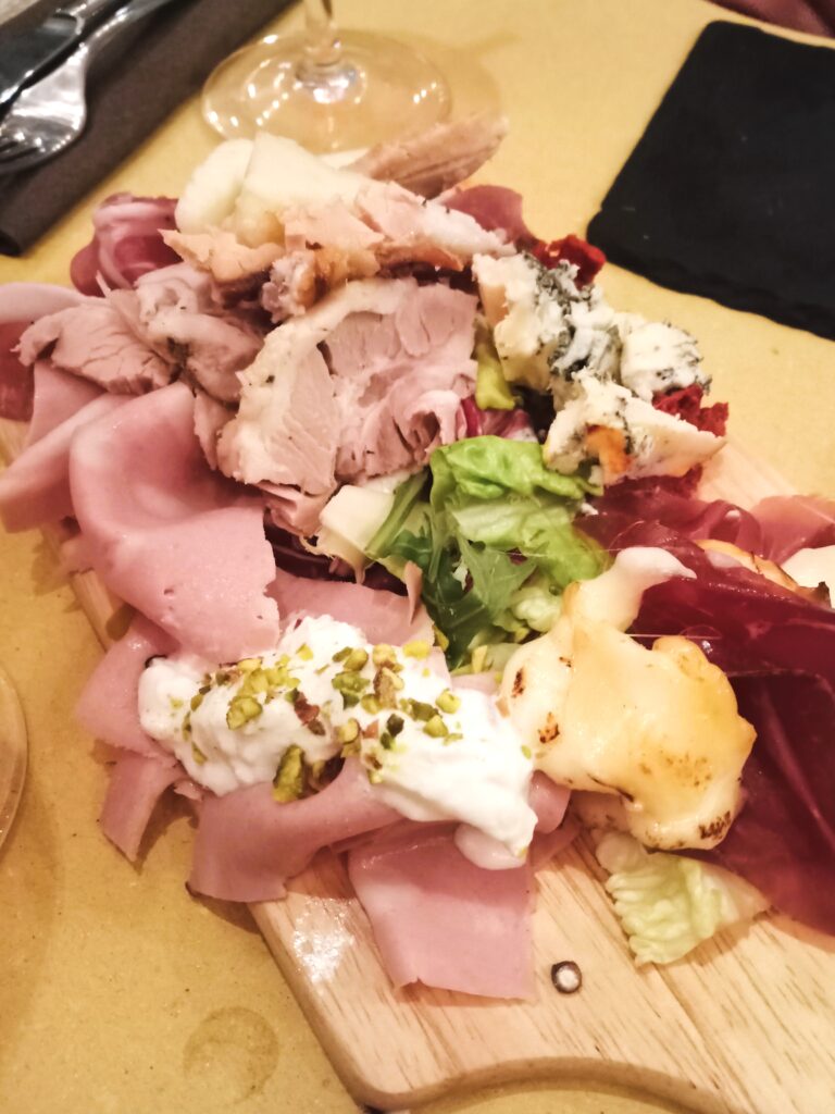 A wooden board with assorted meats, cheeses, and lettuce captures the essence of aperitivo time in Rome. A glass of wine is partly visible in the background, adding to the ambiance.