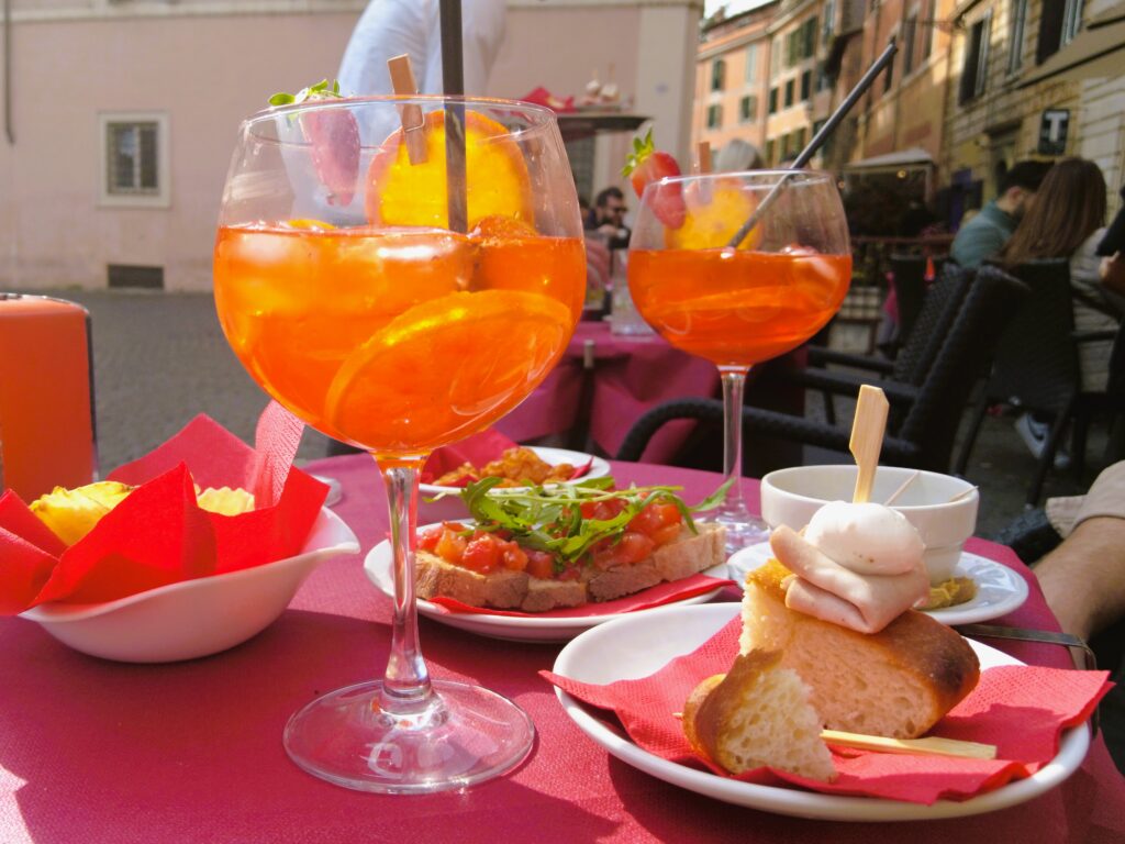 Two glasses of orange cocktails with appetizers sit on an outdoor table, capturing the essence of aperitivo time in Rome under the sunlit cafe's charm.