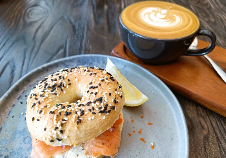 A seeded bagel with salmon and a lemon wedge sits on a plate beside a latte with artful swirls on a wooden table, capturing the essence of one of the best coffee shops in New York City.