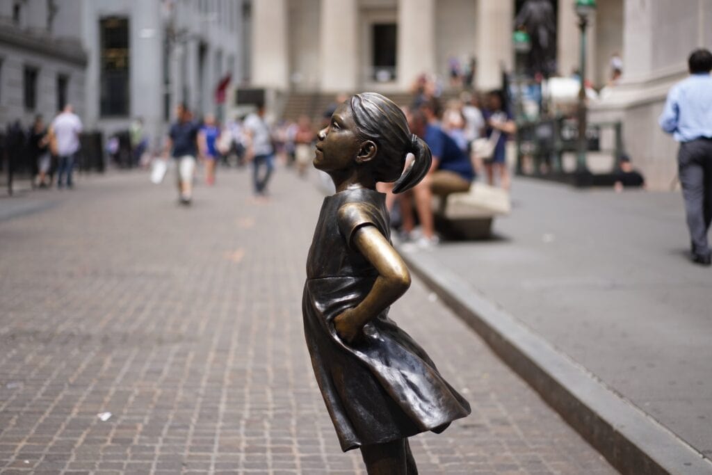 Statue of a girl with hands on hips in a plaza at Battery Park, with people and buildings in the background.