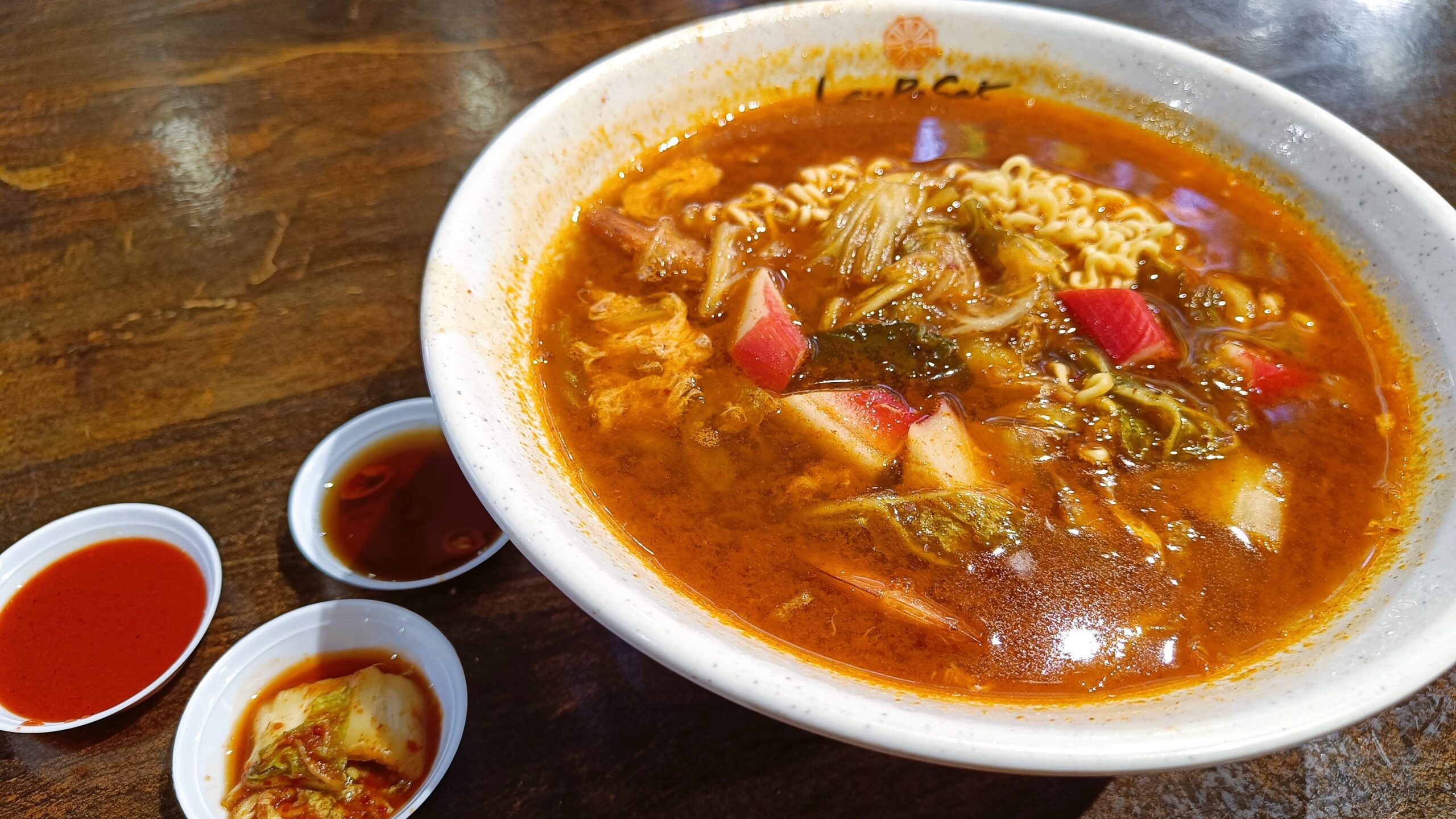 A bowl of spicy ramen with vegetables, accompanied by small dishes of kimchi and red sauce on a wooden table, is a must-try food in Singapore.