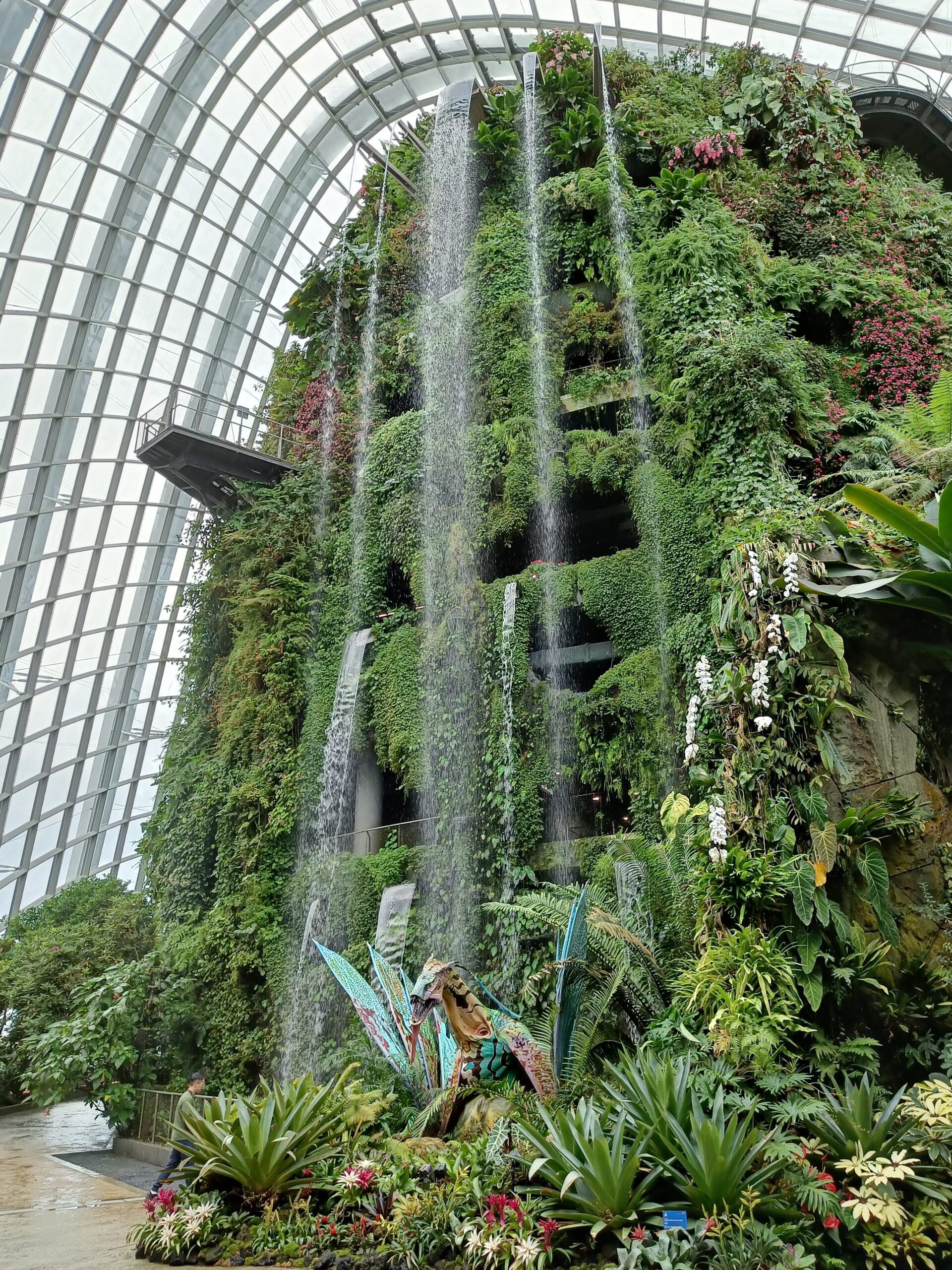 In Singapore's Cloud Forest, a lush indoor waterfall cascades amidst tropical plants, complemented by a statue nestled within the glass-domed conservatory.