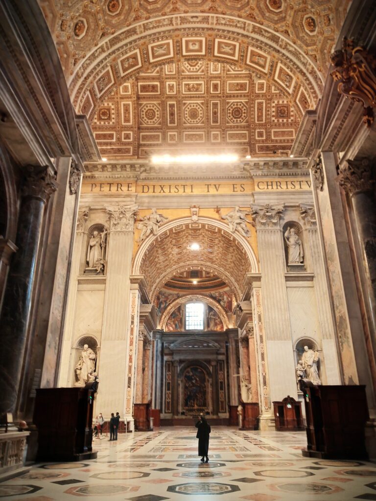 Intricately designed cathedral interior in Vatican City, featuring a detailed ceiling and statues, as a visitor strolls down the aisle. nct