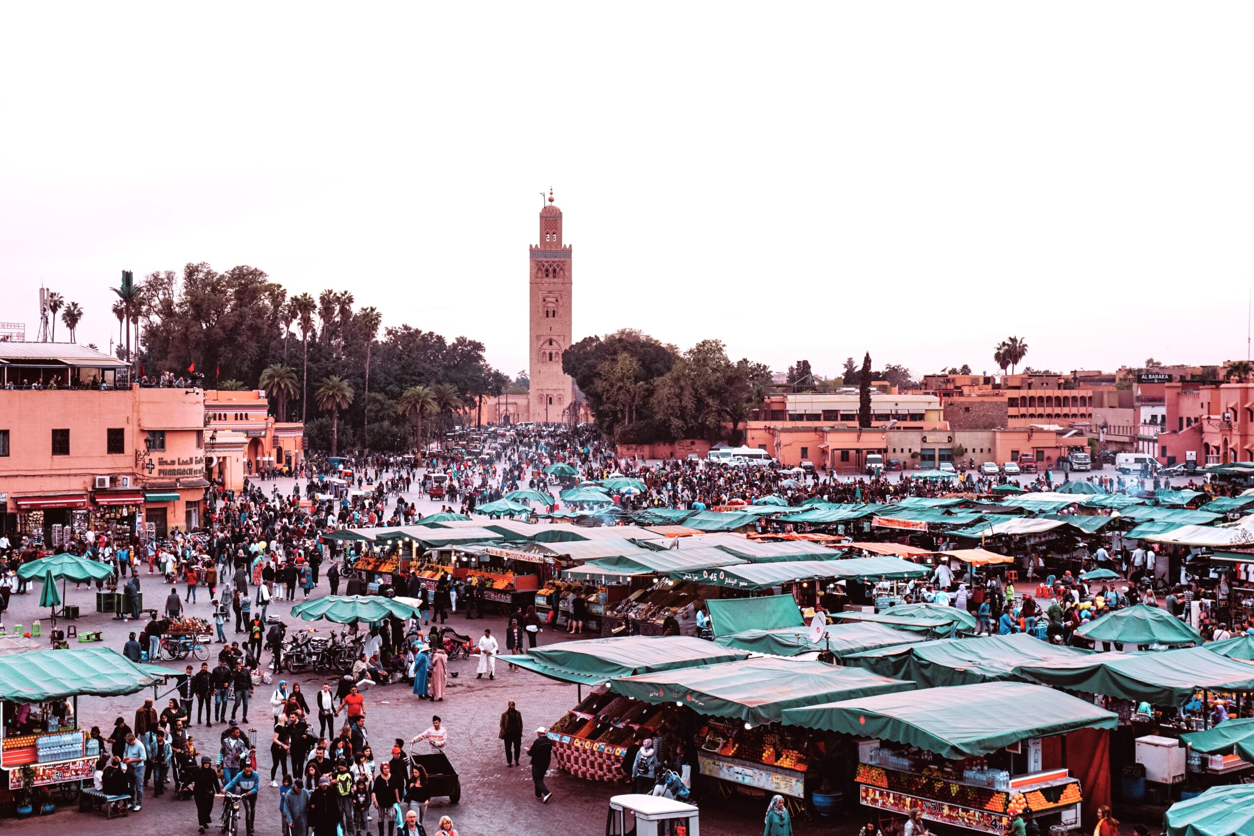 Busy market square in Marrakech with green canopies and a tall mosque tower under a clear sky.