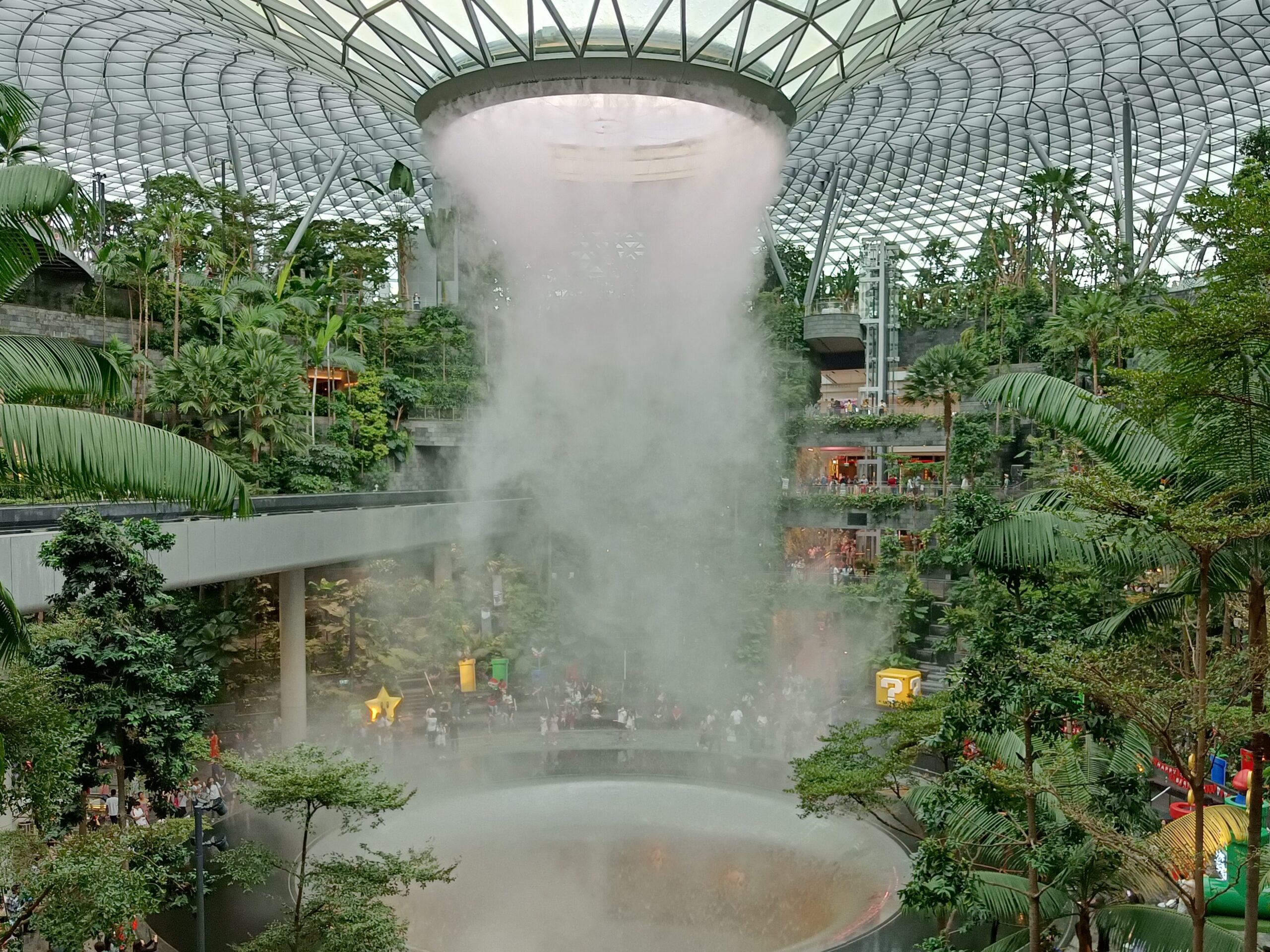 The indoor waterfall, one of the most popular attractions in Singapore, cascades with mist surrounded by lush plants and people under a grand glass dome.