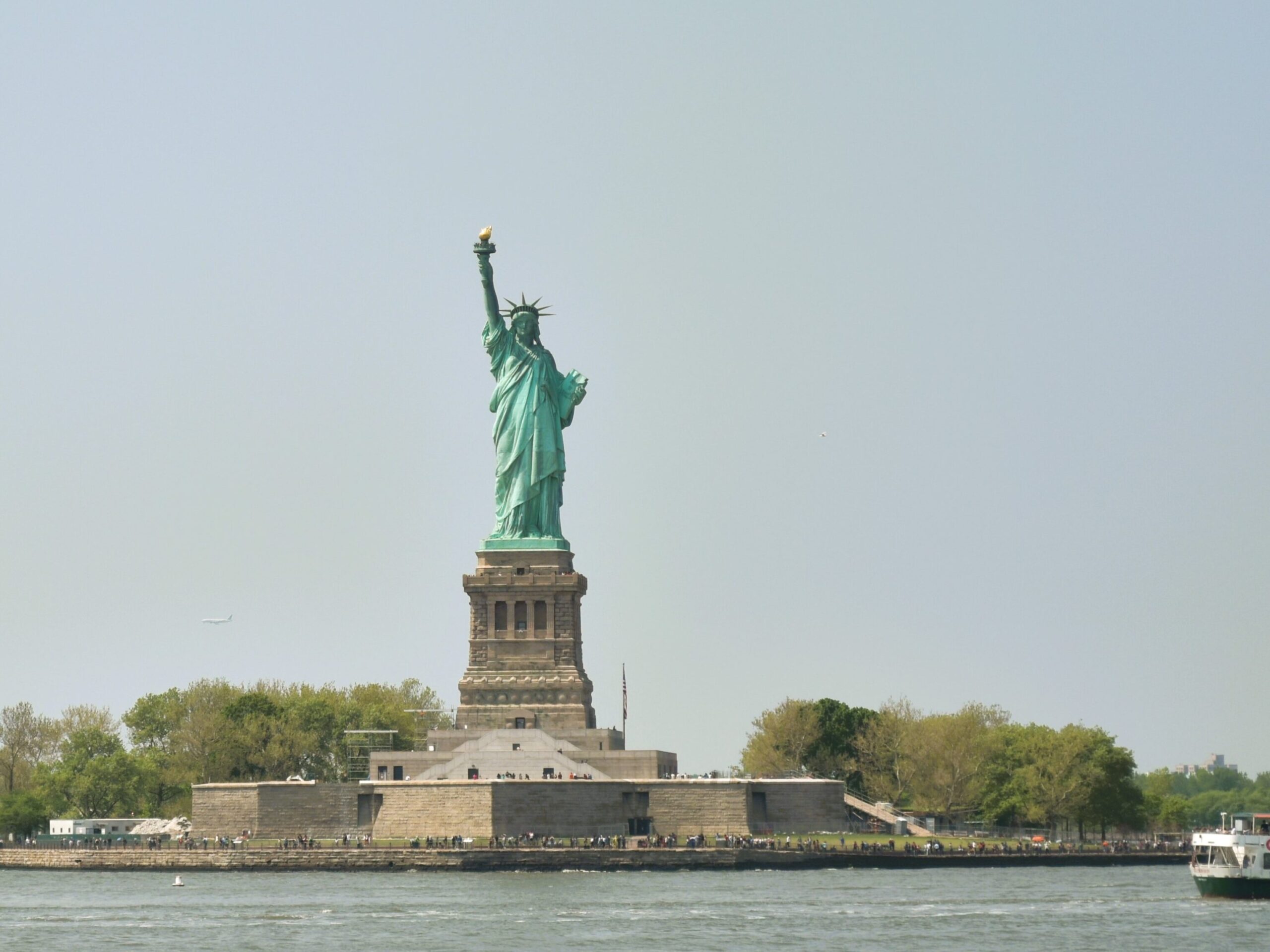 The iconic Statue of Liberty stands majestically on Liberty Island, framed by a clear sky and reflected in the calm water.