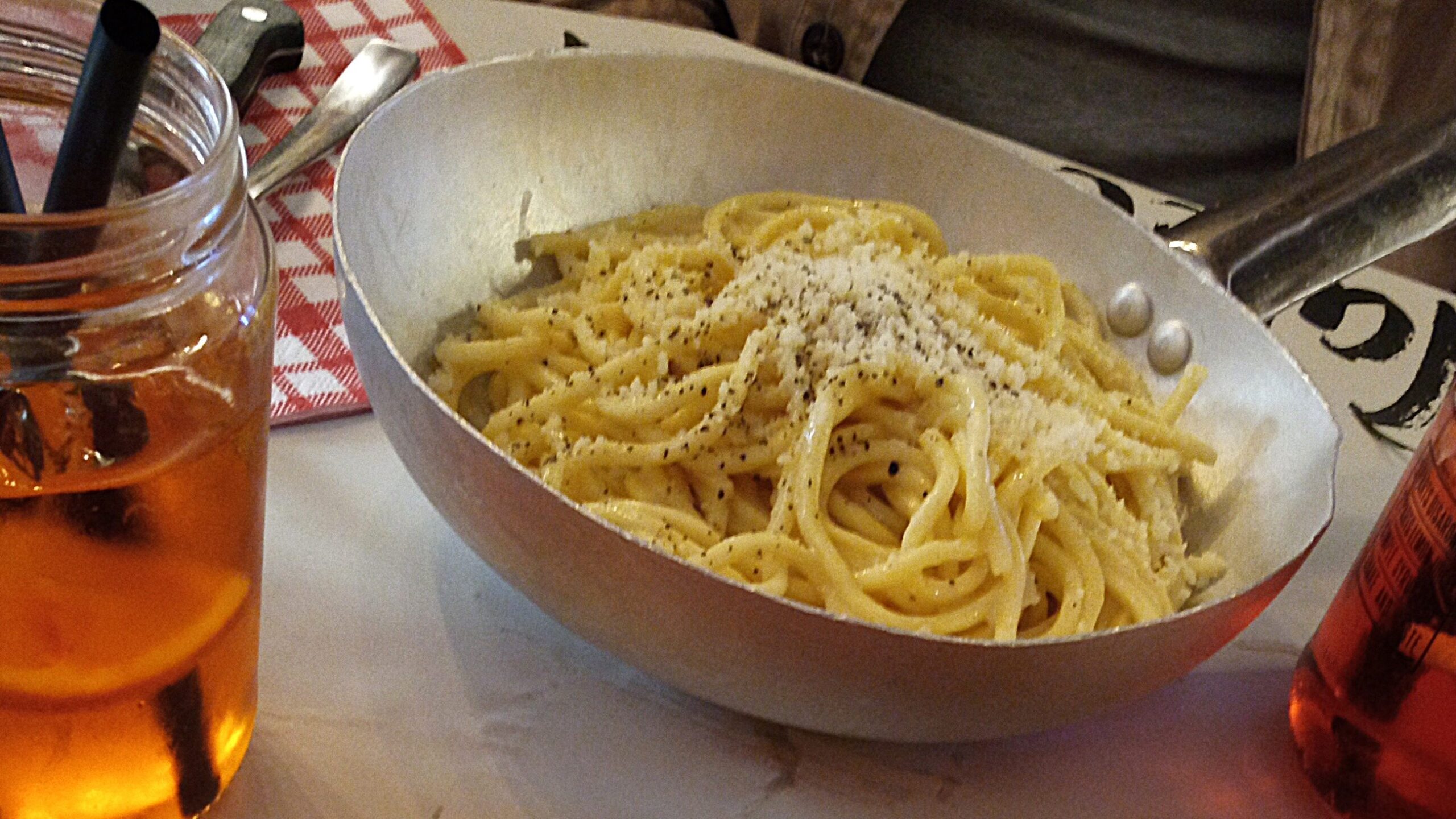 A bowl of spaghetti with cheese and pepper, showcasing the best Italian food, is accompanied by drinks and sliced meat on a rustic wooden table.