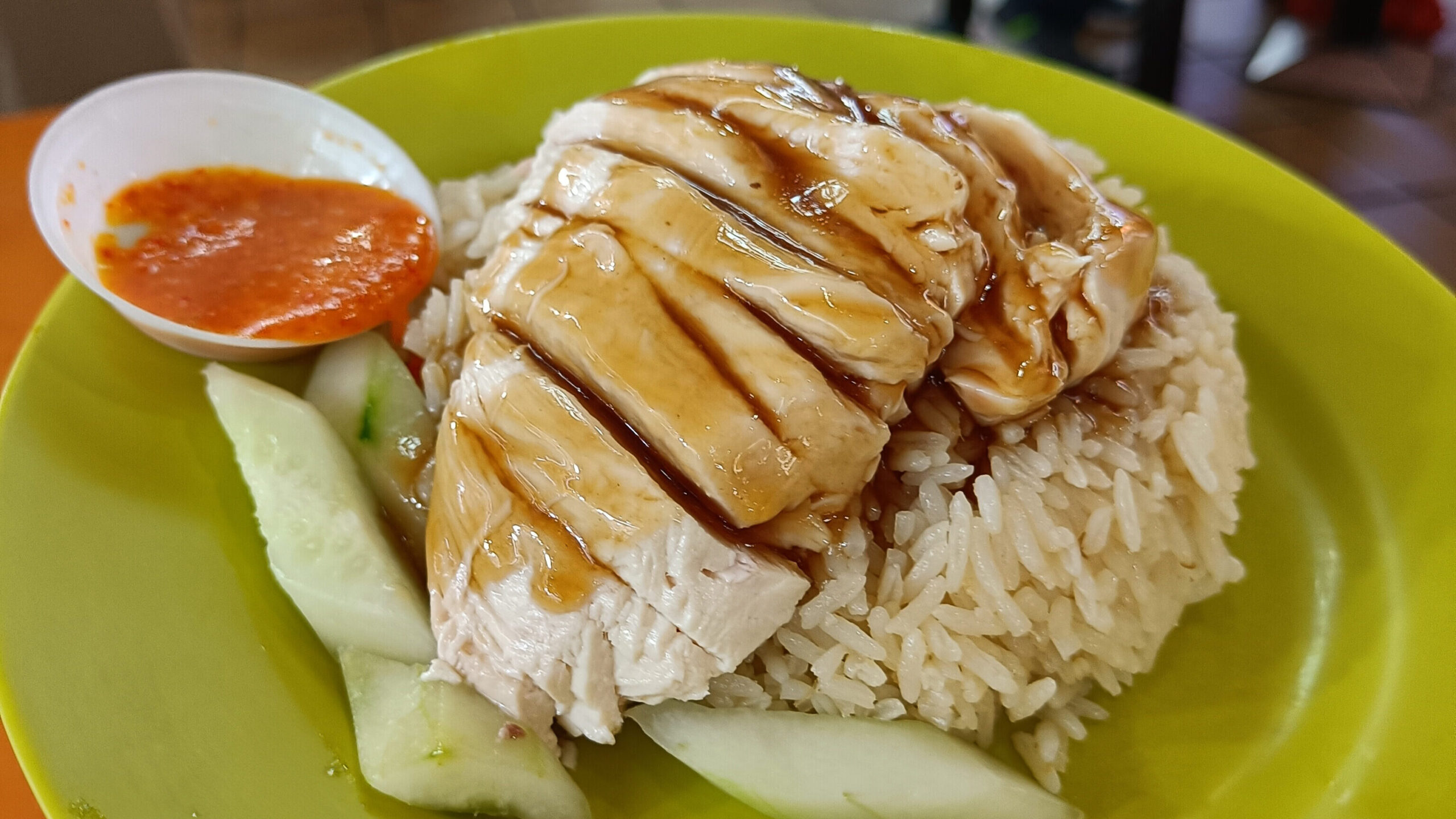 A plate of chicken rice, a must-try food in Singapore, adorned with cucumber slices and chili sauce, served on a bustling food court table.