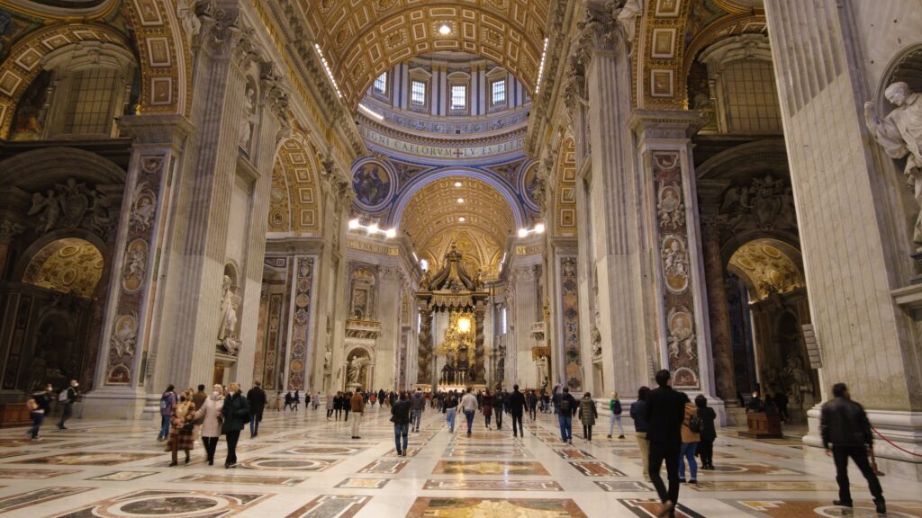 The interior of the grand cathedral in Vatican City boasts ornate columns and a beautifully decorated ceiling, as visitors stroll across the patterned floor, soaking in the majestic ambiance of this iconic Roman site. NCT