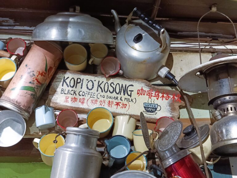 Vintage pots, kettles, and cups adorn a wall with a sign for "Kopi 'O' Kosong" in multiple languages, capturing the rich coffee culture of Singapore.