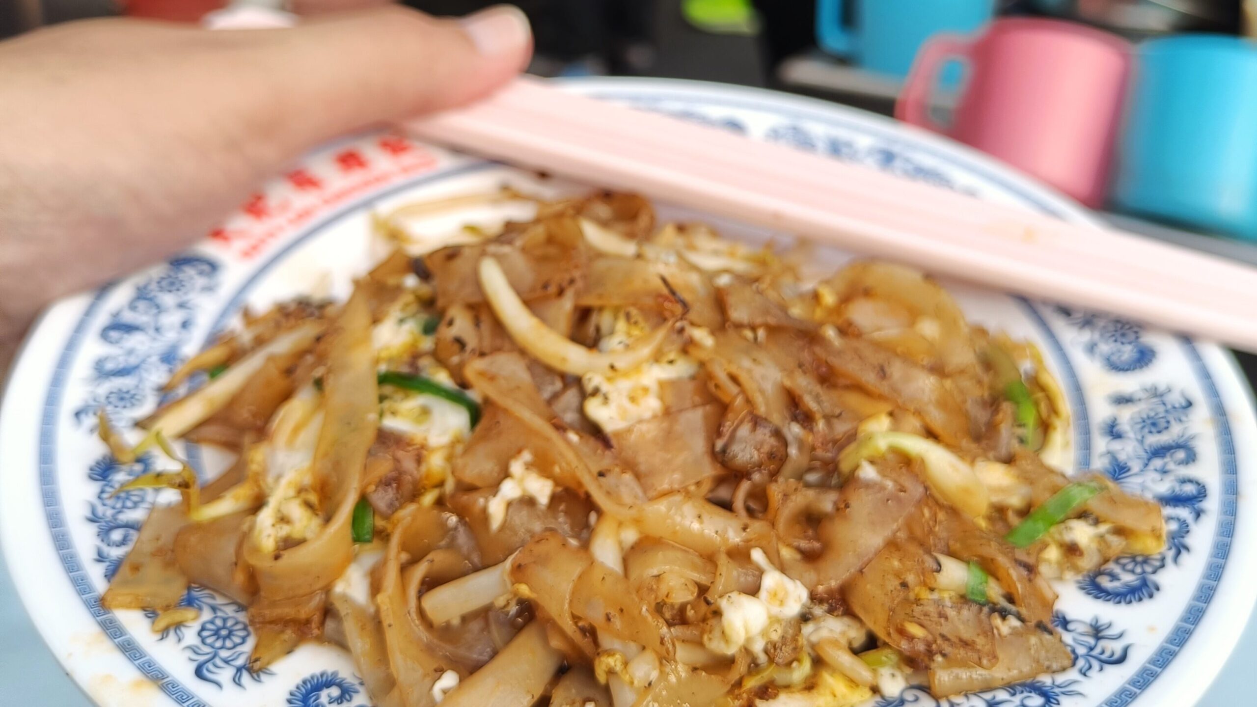 Close-up of a hand holding a plate of stir-fried noodles with chopsticks, showcasing Singapore's must-try food at a bustling street food stall in the blurry background.