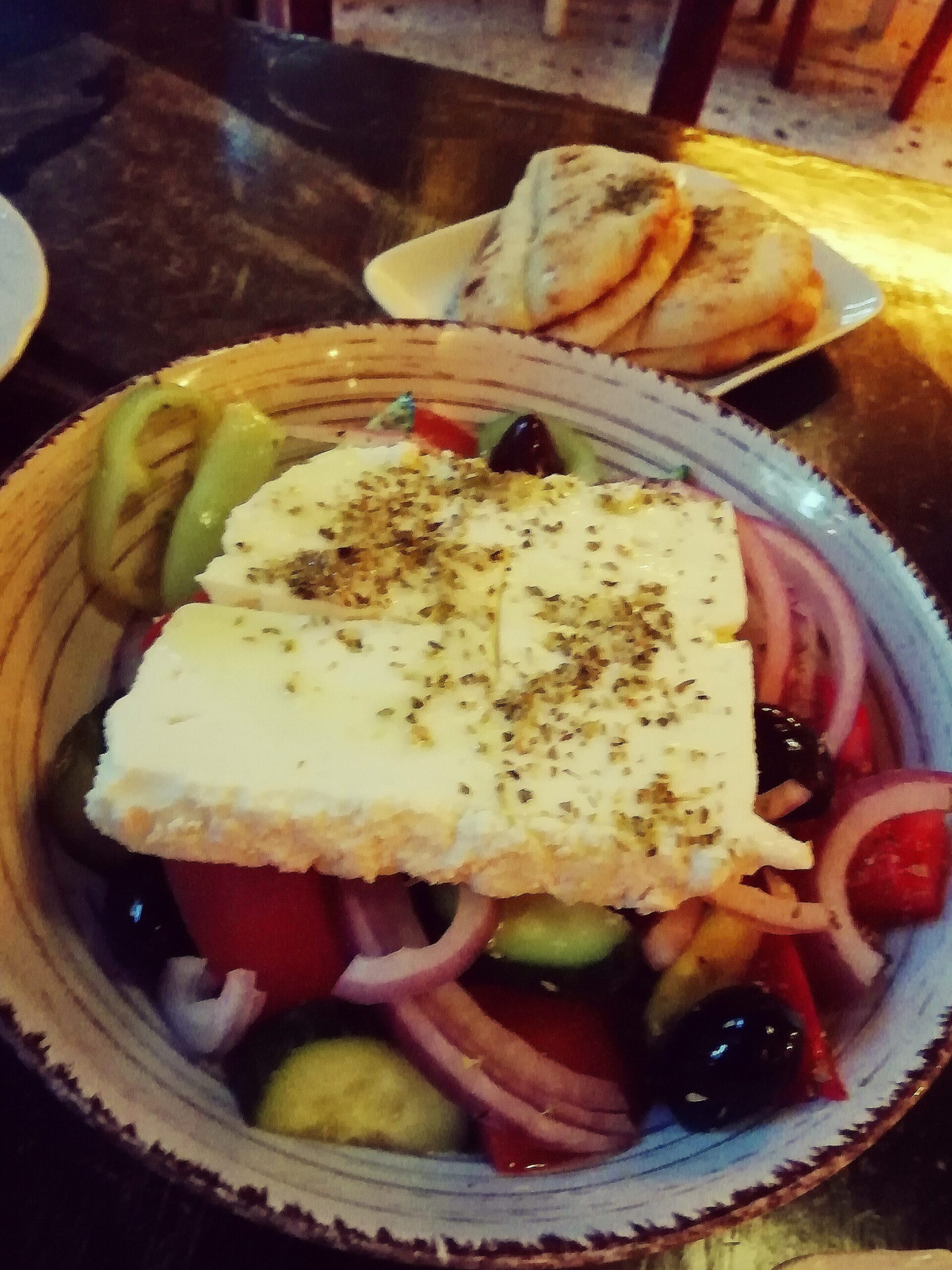 A Greek salad topped with feta, oregano, and olives, served beside pita bread on a dark table—the taste of Athens in one bite. Discover why it's a favorite at some of the best restaurants around.