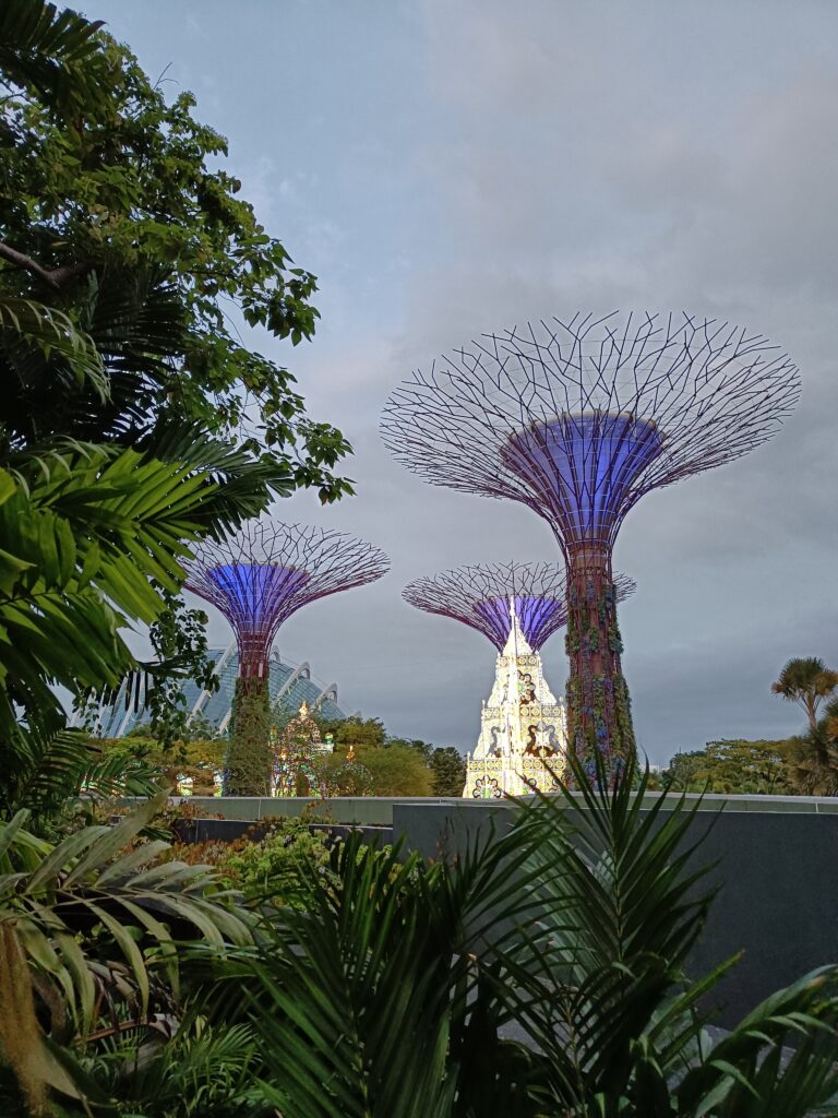 Futuristic tree structures with purple lighting stand majestically against a cloudy sky, surrounded by lush greenery. These captivating installations are among the popular attractions in Singapore, drawing visitors from all over to experience their unique beauty.